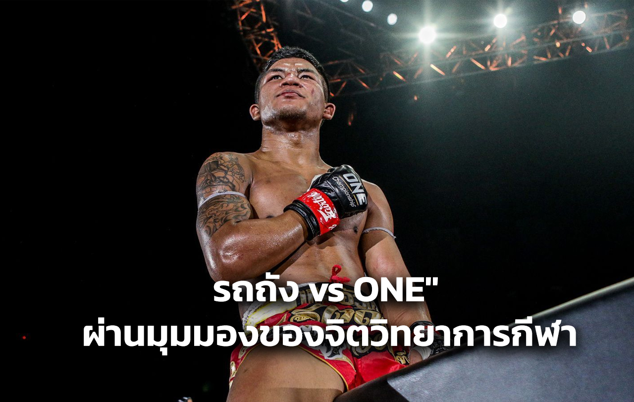Male Muay Thai fighter standing in a ring, sweat on his chest, red shorts and gloves, arena lights above him.